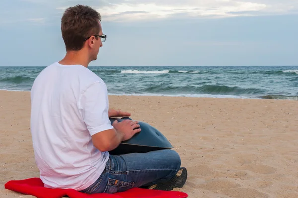 Junge stylische Kerl sitzt am Sandstrand und spielt Handpfanne oder hängen mit Meer auf dem Hintergrund. Der Hang ist ein traditionelles ethnisches Trommel-Musikinstrument — Stockfoto