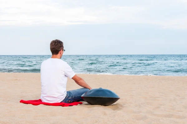 Stylové mladík sedící na písečné pláži poblíž handpan nebo zablokuje s mořem v pozadí. Pověste je tradiční etnické bicí hudební nástroj — Stock fotografie