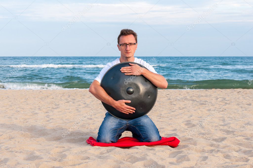 Young stylish guy sitting on the sand beach and holding a handpan or hang with sea On Background. The Hang is traditional ethnic drum musical instrument