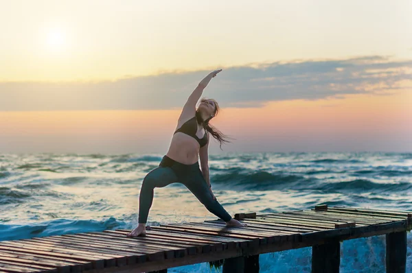 Woman practicing Warrior yoga pose outdoors over ocean or sea and sunset sky background. — Stock Photo, Image