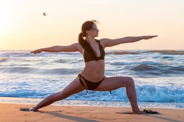 Jonge vrouw ontspannen op het strand, mediteren in warrior asana, bij zonsondergang of zonsopgang en zee of oceaan achtergrond, close-up — Stockfoto