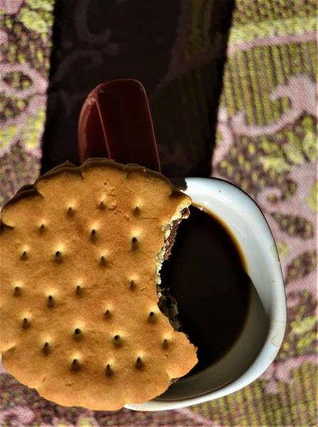 Coffee and double decker biscuit with chocolate. Biscuit with chocolate between two layers bitten on one side, biscuit resting on a red cup. There is one filter of coffee in the cup.