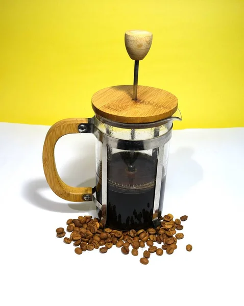 French press coffee maker on the white ground. Arabic coffee beans bottom of the french press and cooked caffe inside the glass side of the press and wooded details with dull yellow background.
