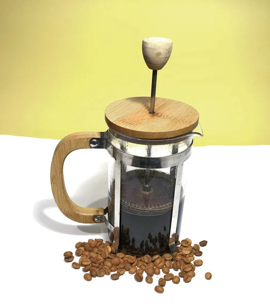 French press coffee maker on the white ground. Arabic coffee beans bottom of the french press and cooked caffe inside the glass side of the press and wooded details with dull yellow background.