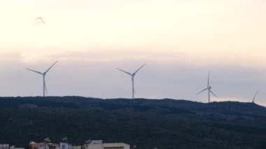 Yavaş çekim videosu. Çok bulutlu bir arka planda hasat yapmadan önce küçük tepelerin ve tarım alanının tepesindeki rüzgar türbini kuleleri.