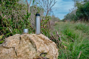 Kafe kupası ve ısı yalıtımlı termos paslanmaz çelikten yapılmış kayayı ormanda vahşi doğada yürürken fotoğraf çekerek kaplıyor.