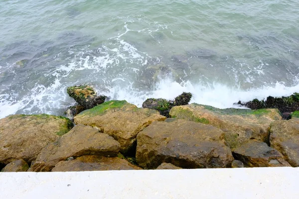 Turquoise ocean and sea water. Orange and yellow stones and rocks are mossed with green. Waves are splashing to rocks during overcast and closed weather.