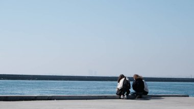 Hindi. İstanbul 04.03.2021. İstanbul Boğazı ve güneşli bir gün. Küçük kızlar Kadıköy kıyısında beton limanda oturur ve dalgakıran arka planına sahip marmarmara denizine bakarlar..