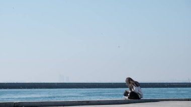 Hindi. İstanbul 04.03.2021. İstanbul Boğazı ve güneşli bir gün. Küçük kız Kadıköy kıyısında beton limanda oturuyor ve dalgakıran arka planına sahip marmarmara denizine bakıyor..