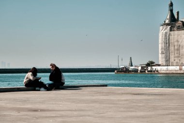 Hindi. İstanbul 04.03.2021.İstanbul Boğazı, güneşli bir gün. Kadıköy kıyısında beton limanda oturan iki genç kız dalgakıran ve sadarpaşa tren istasyonunun arka planında bulunan marmarmara denizine bakıyorlar..