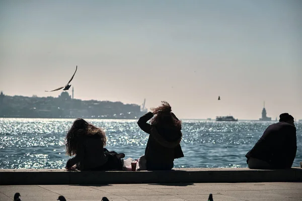 トルコのイスタンブール04 2021 女の子は胸壁の海岸に座っている 巻き毛の女性と彼女の髪の波風からカモメ 船やフェリーの背景と海の朝と太陽の反射中 — ストック写真