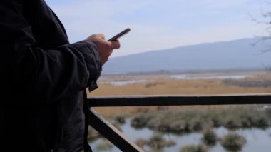 Kuş izleme kulesinde tek bir adam cep telefonuyla kameranın önünde ve karacabey sel ovasının (longoz ormani) arka planında ve bataklık doğası panoramasında oynuyor.