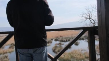 Bir adam kuş izleme kulesinden Karacabey taşkın ovasına bakıyor ve 19. salgından dolayı yeşil tıbbi maske takıyor ve sonra dağ, bataklık ve su olarak görülen doğa sahnesine geri dönüyor..