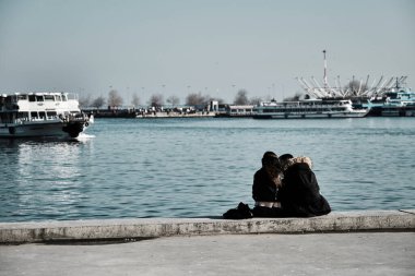 Türkiye istanbul 04.03.2021. Güneşli bir günde, Kadıköy 'de birbirine sarılıp boğaza yaslanan iki genç çift ve sevgili yolcu feribotu ve turkuaz su arka planıyla İstanbul' da oturuyorlar..