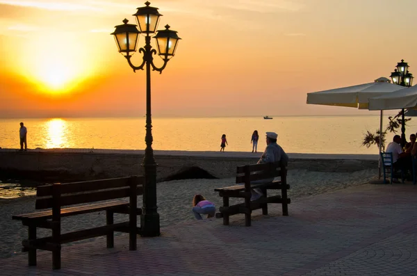 Old man enjoys sunset at the bench by the sea while grandchildren are playing in the sand