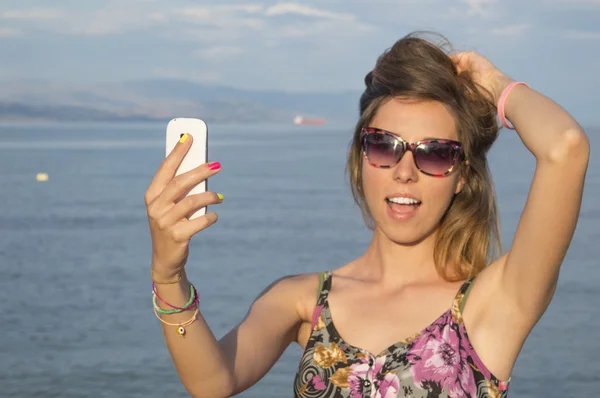 En telefon i unge piger hånd tager selfie på havet iført sol - Stock-foto