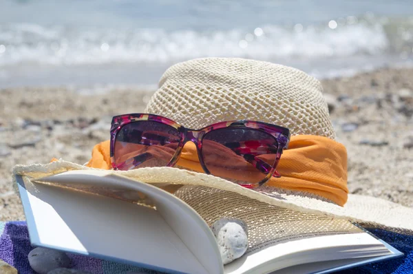 Chapéu de palha óculos de sol e um livro na praia com mar em backgou — Fotografia de Stock