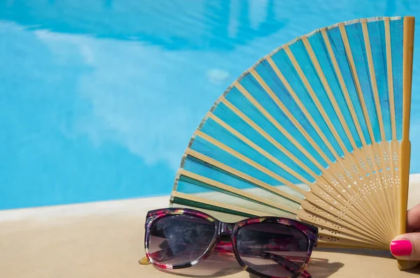 Hand-fan in girls hand next to sunglasses with clear blue water — Stock Photo, Image