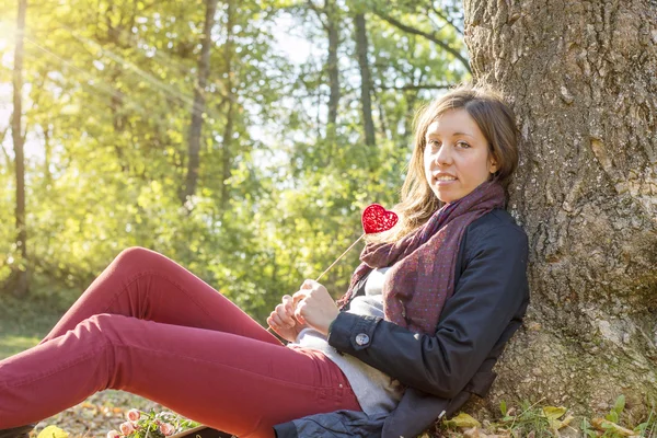 Hermosa chica sosteniendo el corazón rojo en un palo mientras está sentado en el —  Fotos de Stock