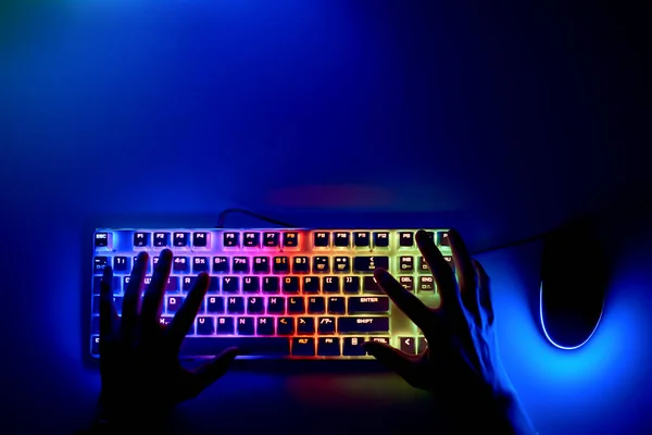 Hands on the keyboard in the first person. A hacker is sitting at a computer in a dark room. The concept of cybercrime. Keyboard and mouse with RGB light and human hands