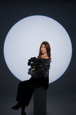 Siyah bir elbise giyen ve elinde siyah çiçekler tutan göz alıcı bir kadının zarif stüdyo portresi dramatik dairesel bir sahne ışığı altında fotoğraflandı..