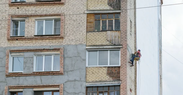 Строитель-альпинист висит на веревке с крыши и штукатурщики кирпичной стены многоэтажного жилого дома — стоковое фото