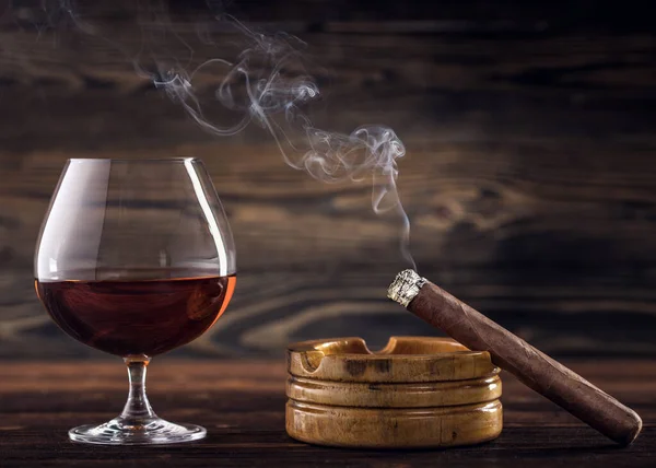 Cognac and cigar. Glass of cognac or brandy with cigars from Cuba Havana and ashtray on natural wooden background. Glass of whiskey with smoke cigar. Alcohol drink on Bar counter in the restaurant.