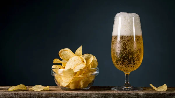 Beer and Potatoes Chips. Glass of blonde beer with a large head of foam. Chips in glass bowl good for snack on natural wooden table. Good for beer festival, pub, restaurant advertising. Food and Drink