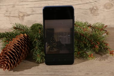 Çalışan ekranlı akıllı telefon ve modern çerçevesiz tasarım, dikey olarak köknar dalları ve çam kozalaklarıyla ahşap bir arka planda. Oda içi tasarım ekranında çalışan akıllı telefon.