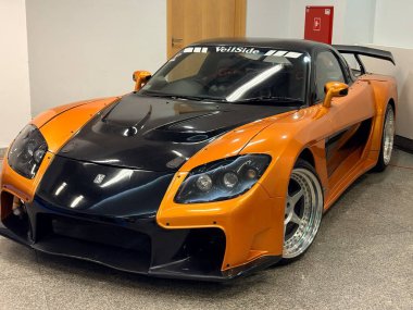 01.01.2026 Polonya, Varşova otomobil sergisi, Turuncu ve siyah Mazda RX-7 Veilside Fortune vücut kiti kapalı bir araba fuarında, sürüklenme ve performans için özel Japon spor arabası.