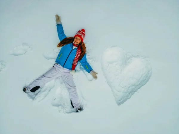 A heart. A woman lies in the snow next to a drawn heart. Aerial view.