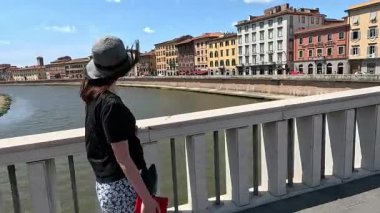 Şapkalı bir turist köprü boyunca yürür ve Pisa 'daki evlere bakar..