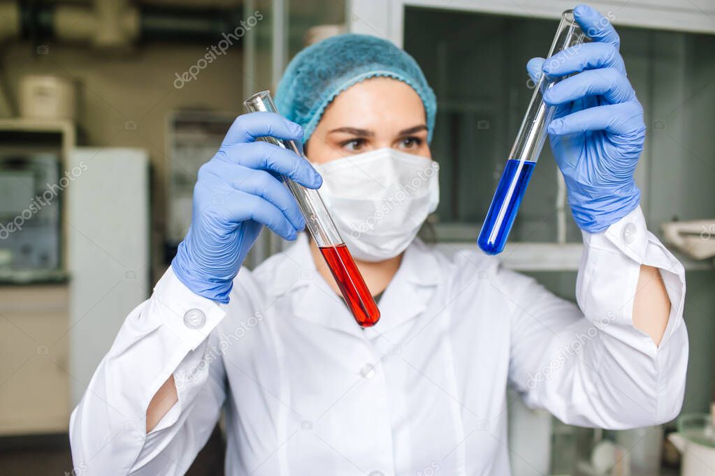Young female scientist in medical cap and protective mask are doing investigations with test tubes red and blue solution in laboratory.