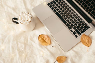 Laptop, sarı sonbahar yaprakları, şekerli bir fincan kakao, parlak ışıklar, beyaz yatakta mum. Ev konseptinde çalış. Sonbahar, sonbahar, kış konsepti. Düz konum, üst görünüm, kopyalama alanı.