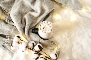 Bir fincan kakao, marşmelov, atkı, pamuk, beyaz çarşafta parlayan ışıklar. Noel, kış, sonbahar yeni yıl konsepti. Düz konum, üst görünüm, kopyalama alanı.