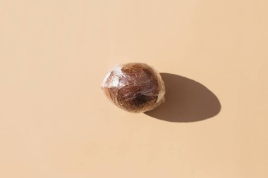 Bej arka planda polietilen plastiğe sarılmış hindistan cevizi. Asgari çevresel konsept. Meyve konsepti. Düz düz, üst görünüm