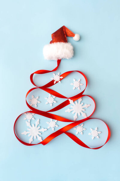 Творческая елка из снежинки, красной ленты и шапки Санта-Клауса на светло-голубом фоне. Рождество, зима, новый год. Плоский, вид сверху