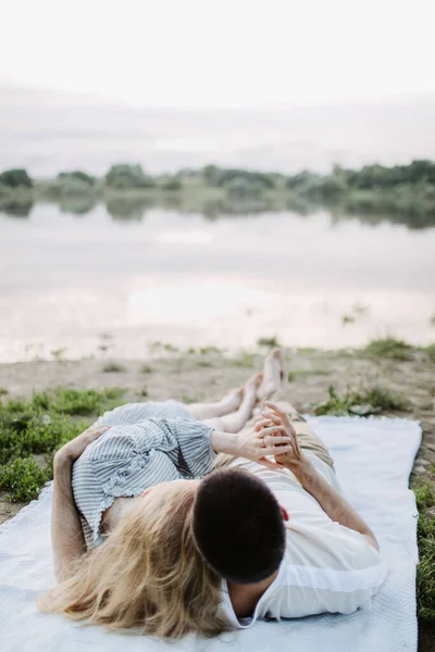 Giovane uomo abbraccia e tiene la mano di una bella donna. Gli amanti giacciono su una coperta vicino al fiume. — Foto Stock