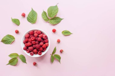 Beyaz kasede taze ahududu ve pastel pembe arka planda yeşil yapraklar. Düz konum, üst görünüm, kopyalama alanı