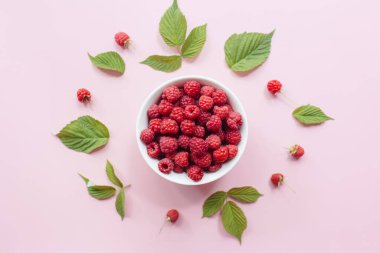 Beyaz kasede taze ahududu ve pastel pembe arka planda yeşil yapraklar. Düz yatış, üst görünüm