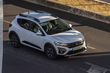 Beyaz Dacia Sandero Stepway SUV, arka planı bulanık asfalt bir yolda sürüyor.