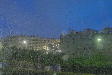 Gün batımında bulanık şehir ışıkları ve binaları olan bir pencereye yağmur damlaları düşüyor.