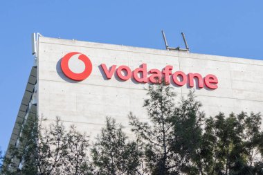 Vodafone logosu ve markası mavi gökyüzünün altında modern bir ofis binasının ön cephesinde sergilenmektedir.
