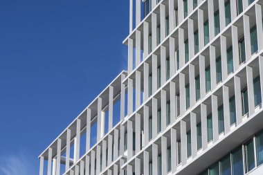 Dikey ve yatay çizgilerle beyaz mimari cephe açık mavi gökyüzüne karşı geometrik desen oluşturuyor