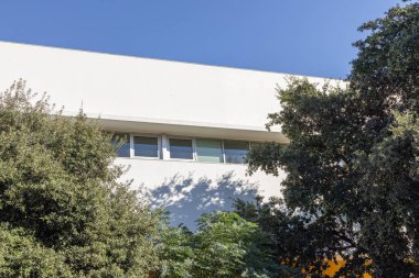 Açık mavi gökyüzünün altında yeşil ağaçlarla çerçevelenmiş beyaz modern bina cephesi