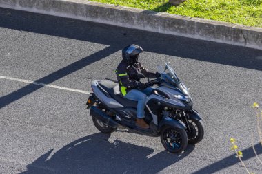 Üç tekerlekli bir yamaha tricity scooter 'la şehir yolunda gezen bir sürücü.