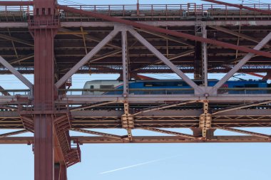 Mavi ve beyaz tren, büyük bir kırmızı asma köprünün alt güvertesinde ilerliyor.