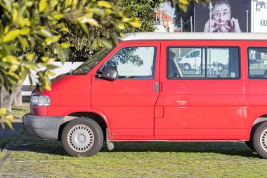Kırmızı minibüs yeşil çimlerin üzerine park edilmiş ve reklam panosu ile arka planda komik bir yüz ifadesi olan bir adam var.