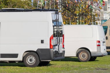 Çatı rafları olan iki beyaz kargo minibüsü ve güneşli yeşil çimlere park edilmiş bir merdiven.