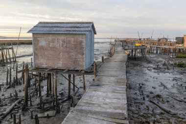 Köhne ahşap balıkçı kulübeleri, çamurlu nehir yatağının üzerindeki iskelelerde ayakta duruyor.